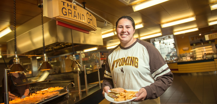 Woman smiling and holding plate of food in front of The Grand Grill at Washakie Dining Center