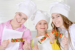 Three girls wearing white chef hats and holding kabob skewers.