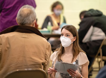 woman in mask with clipboard talking to someone in a busy room