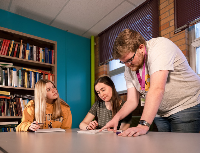 A group of three students look at notes in a library setting.