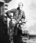 Frewen wearing buckskins leaning on backpack in front of log cabin