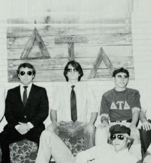 Group photo of the men of Delta Tau Delta all dressed differently, some in suits some in t-shirts, all with black sunglasses on with their flag in the background