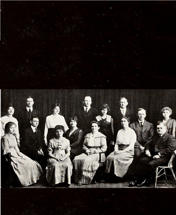 15 people, 9 women and 6 men, posing for the group photo. Women are in dresses men are in suits of the 1910s style.