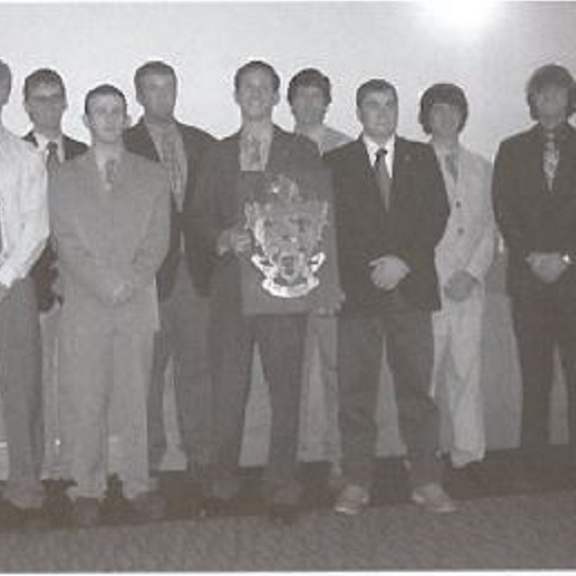 Men of farmhouse, in black and white, holding a plaque with the fraternity crest. All men are in suits.