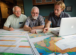 Three men at table examine maps in preparation for an upcoming edition of the scientific journal.