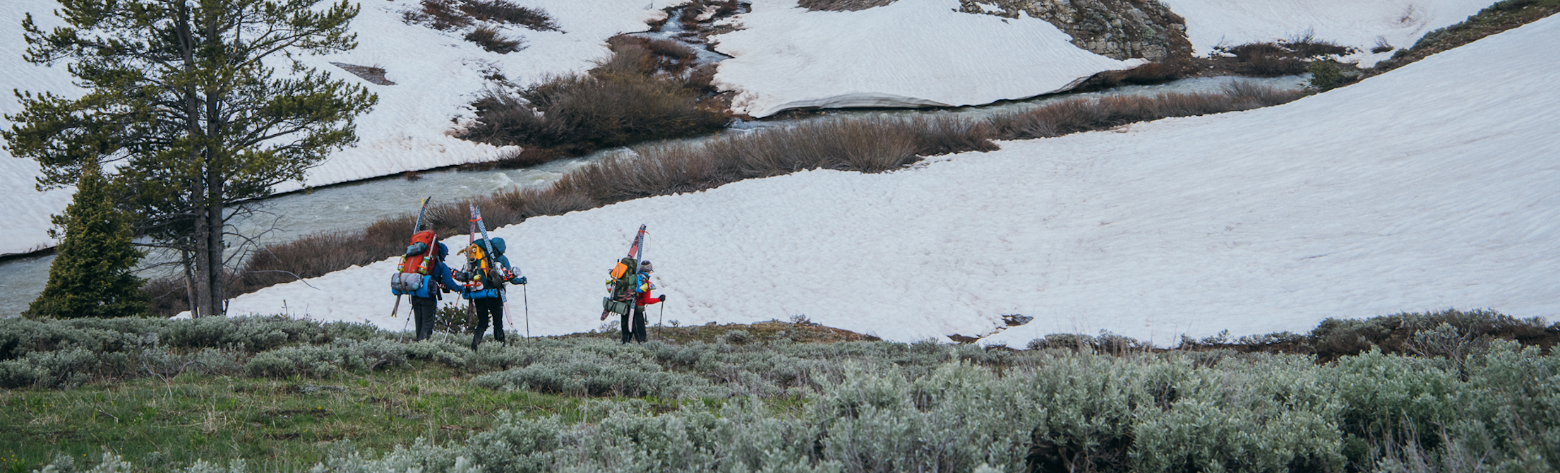Three people carry backpacks with skis on them through the sagebrush and snow along a river.