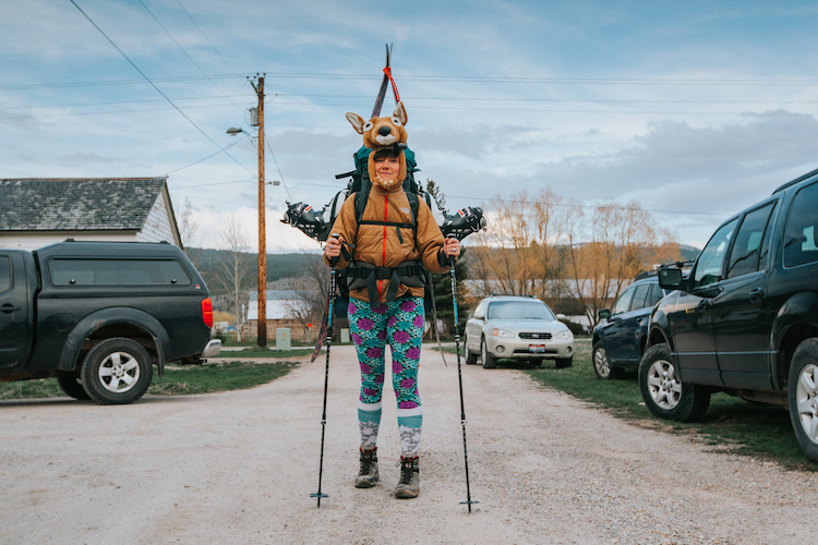 Character from the film Deer 139 wears a backpack with skis attached and a deer costume while standing in a driveway.