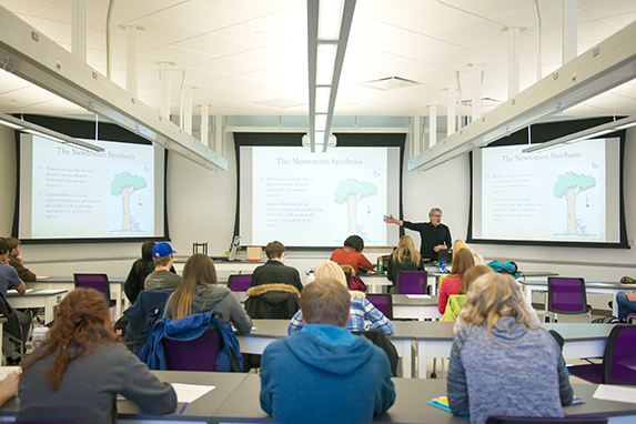 Welcome Picture for Student Material. A picture taken behind students of students sitting in a class with a professor at standing at the front. 