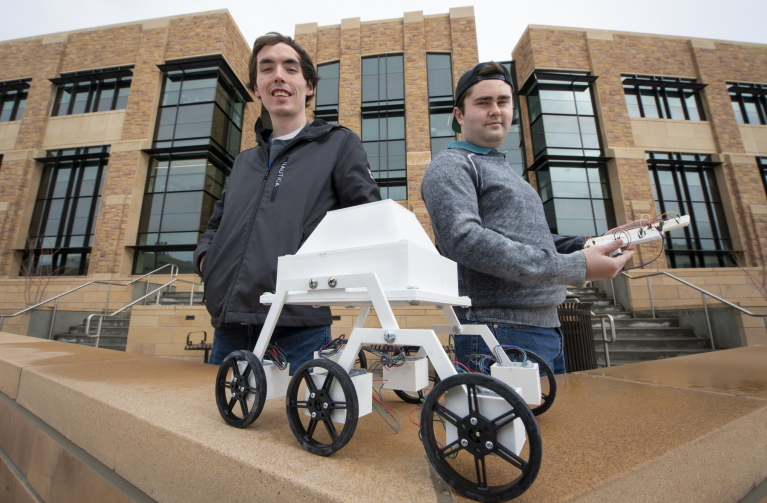 Photo of two ME undergrad students with a robot in front of the EERB