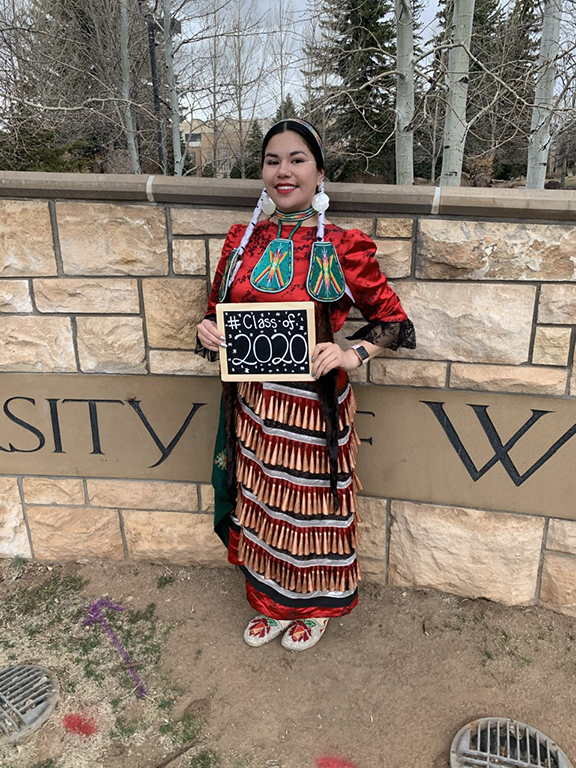 Graduate Christie Wildcat before a university of wyoming sign in native dress holding a plaque that says class of 2020