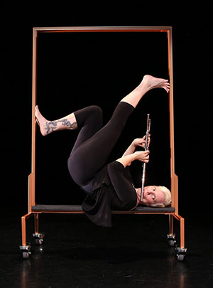 woman playing a flute while posing on a luggage rack on her back with legs in the air