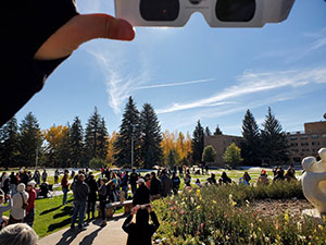 person holding up solar eclipse viewing glasses with a large number of people in the background