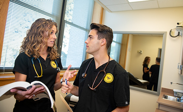 Two nursing students in black scrubs discussing textbook materials in exam room