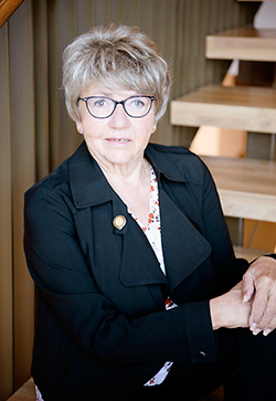 woman with glasses and short feathered hair in black suit with staircase in background