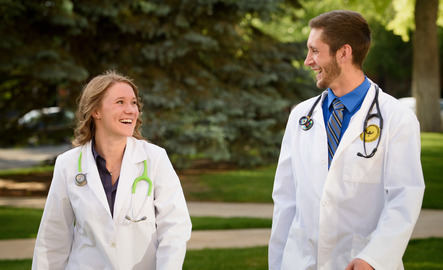 Two students in a nursing program at the University of Wyoming smile at one another as they are walking, wearing lab coats and stethoscopes.