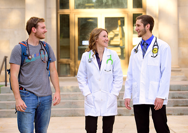 Three students with stethoscopes around their necks visit outside the Health Sciences building