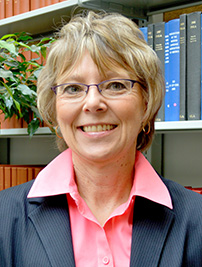 Woman with short blonde hair, pink shirt, grey suit, in front of bookcase