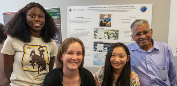 Three students and a professor smile for a photo in front of an informational poster