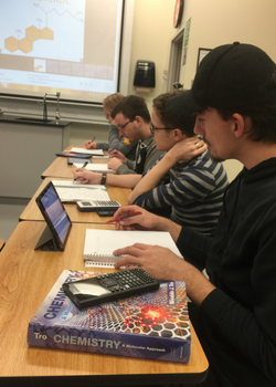 three students at a table with chemistry textbooks, ipads, calculators, and notebooks