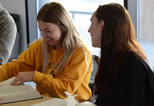 A student in a Wyoming sweatshirt speaking with a UW professor and turning the page of a book