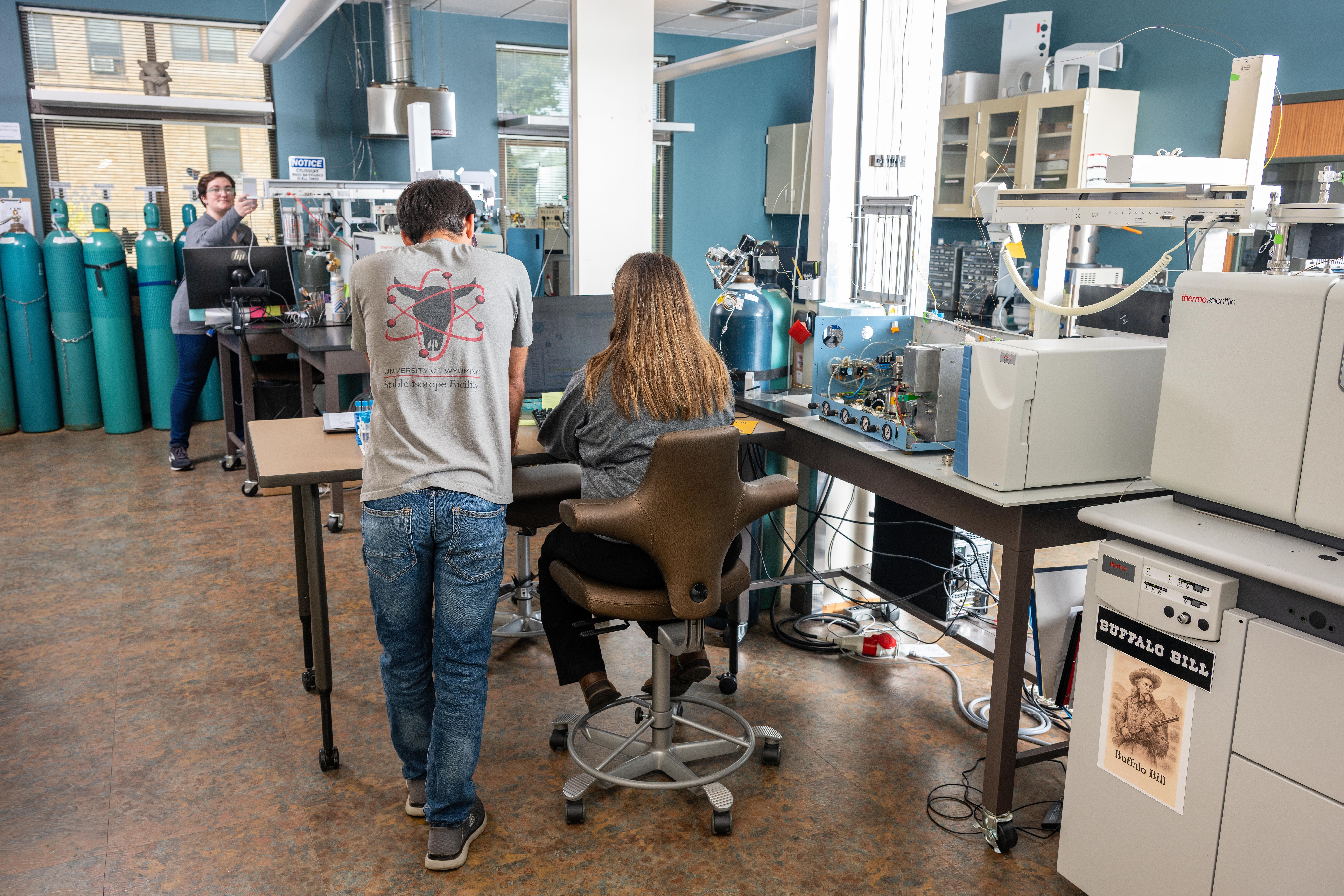 Laboratory technicians working in a lab. Two people are working to gether at a desk and another is in the background working on a scientific instrument.