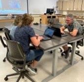 Three MSC students sitting at a table together, working on their laptops with course content projected on a screen in the background