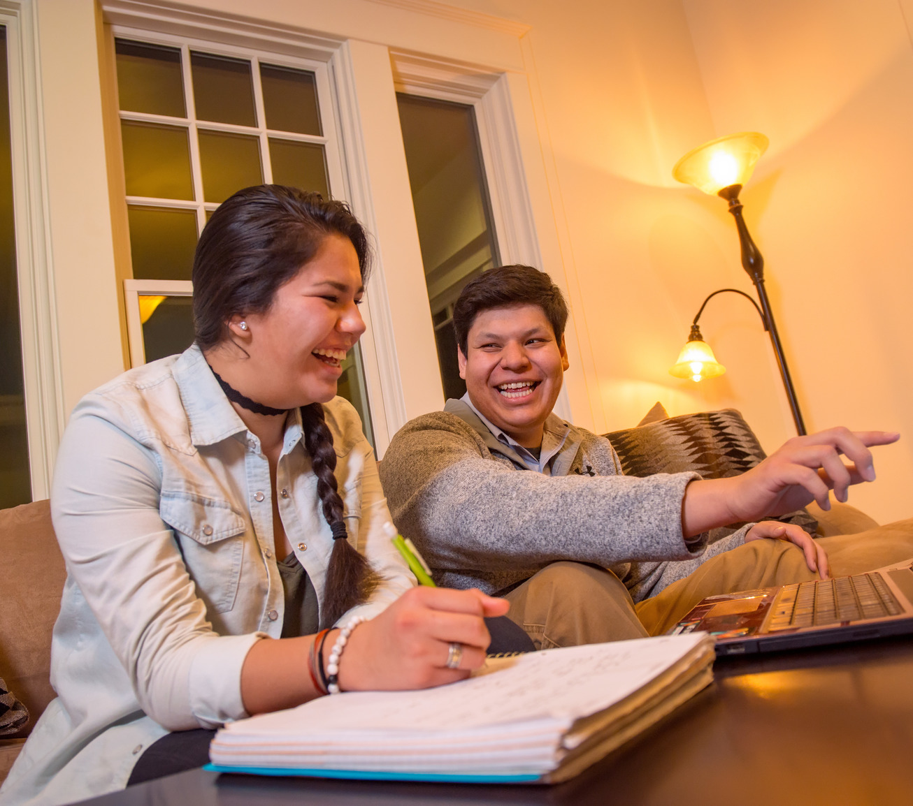 Two students laugh while studying together on a couch.