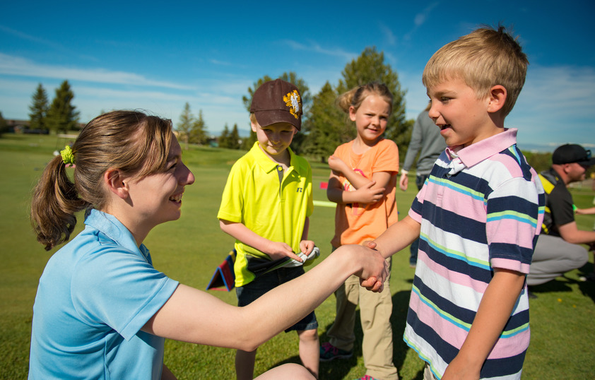 a young woman shakes hands with a child on a green field outside while there are other children in the background