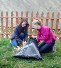 two women crouched on a lawn gathering fallen leaves into a garbage bag