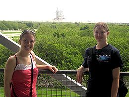 two women standing by railing with vast area of greenery and a rocket gantry behind them