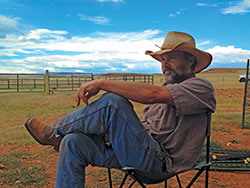 man in cowboy hat sitting outside in a chair, legs crossed, looking into the distance