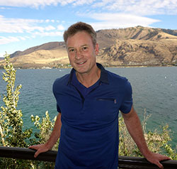 man leaning against a railing with lake and mountain in the background