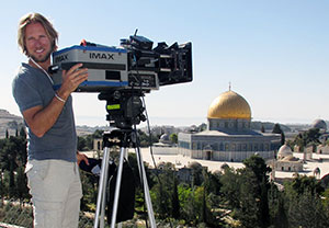 man with a large video camera overlooking a view with ancient buildings