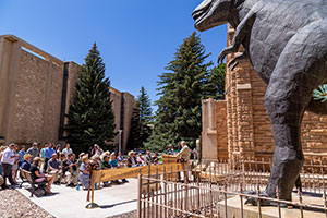 people listening to a person speaking in front of a T-Rex statue