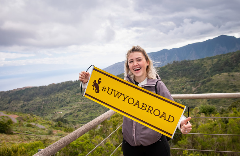 A UW student holding UWYO Abroad banner at a mountainous scenic overlook.