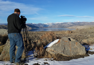 A person person with a camera films a lake and mountains