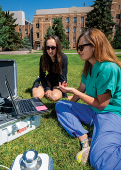 two women sitting in the grass with a laptop