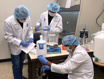 three people in facemasks, head covers and gloves working in a lab