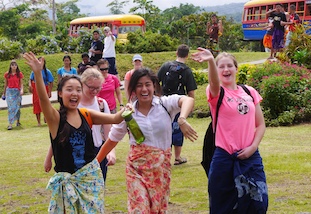 three young women in bright clothing wave happily while crossing a lawn with colorful buses in the background