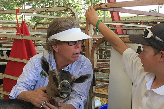 Adult holding a goat visiting with young man.