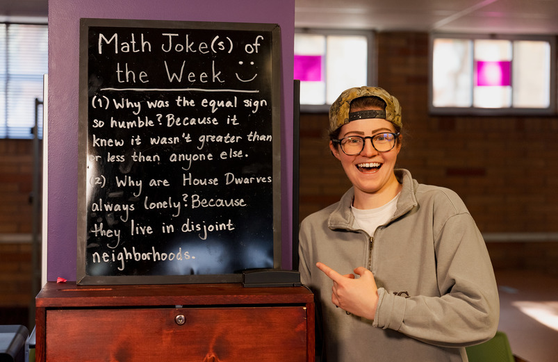 student worker at the math lab standing in front of a chalk board with math jokes written on it