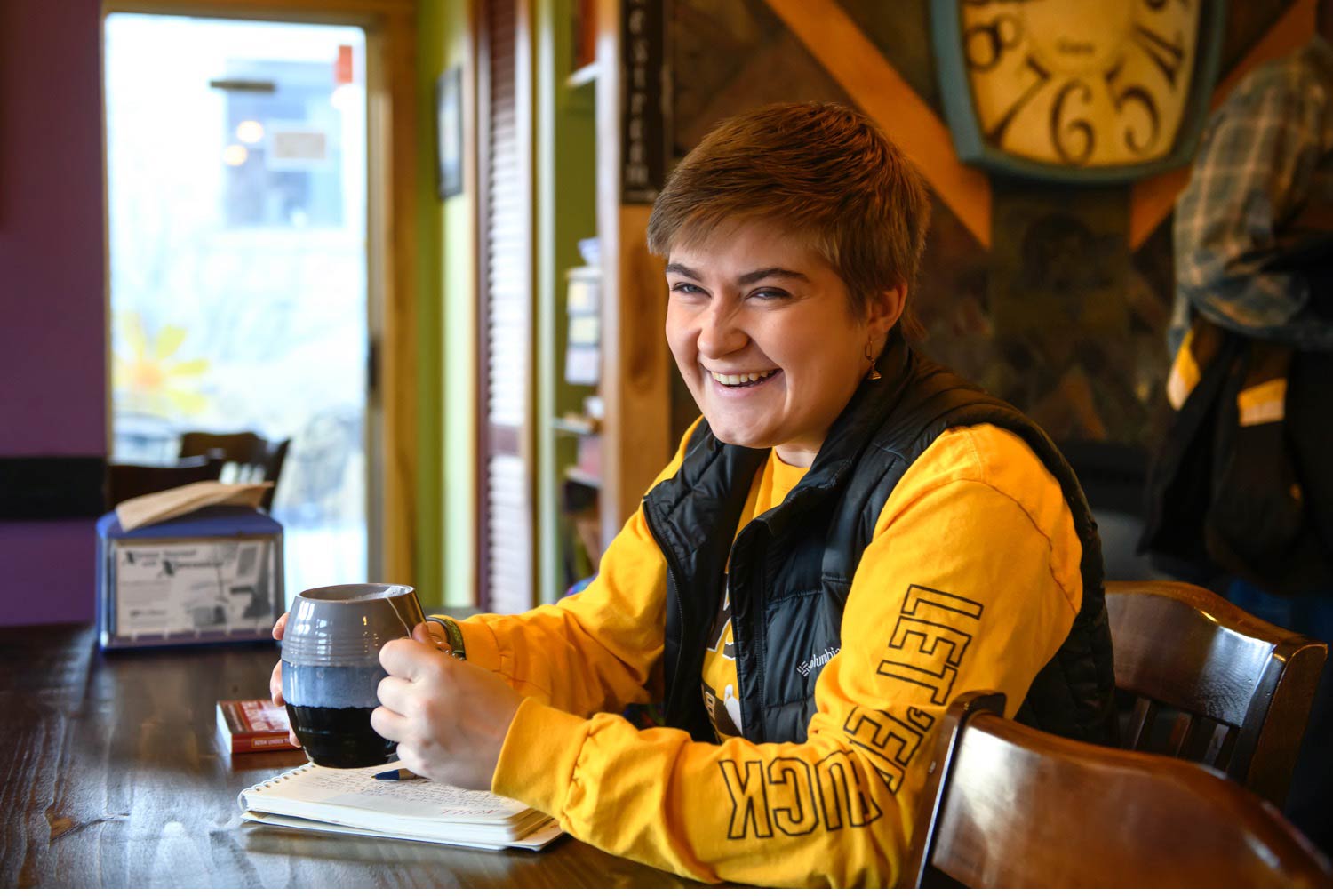 A student sit with some coffee wearing a yellow shirt.