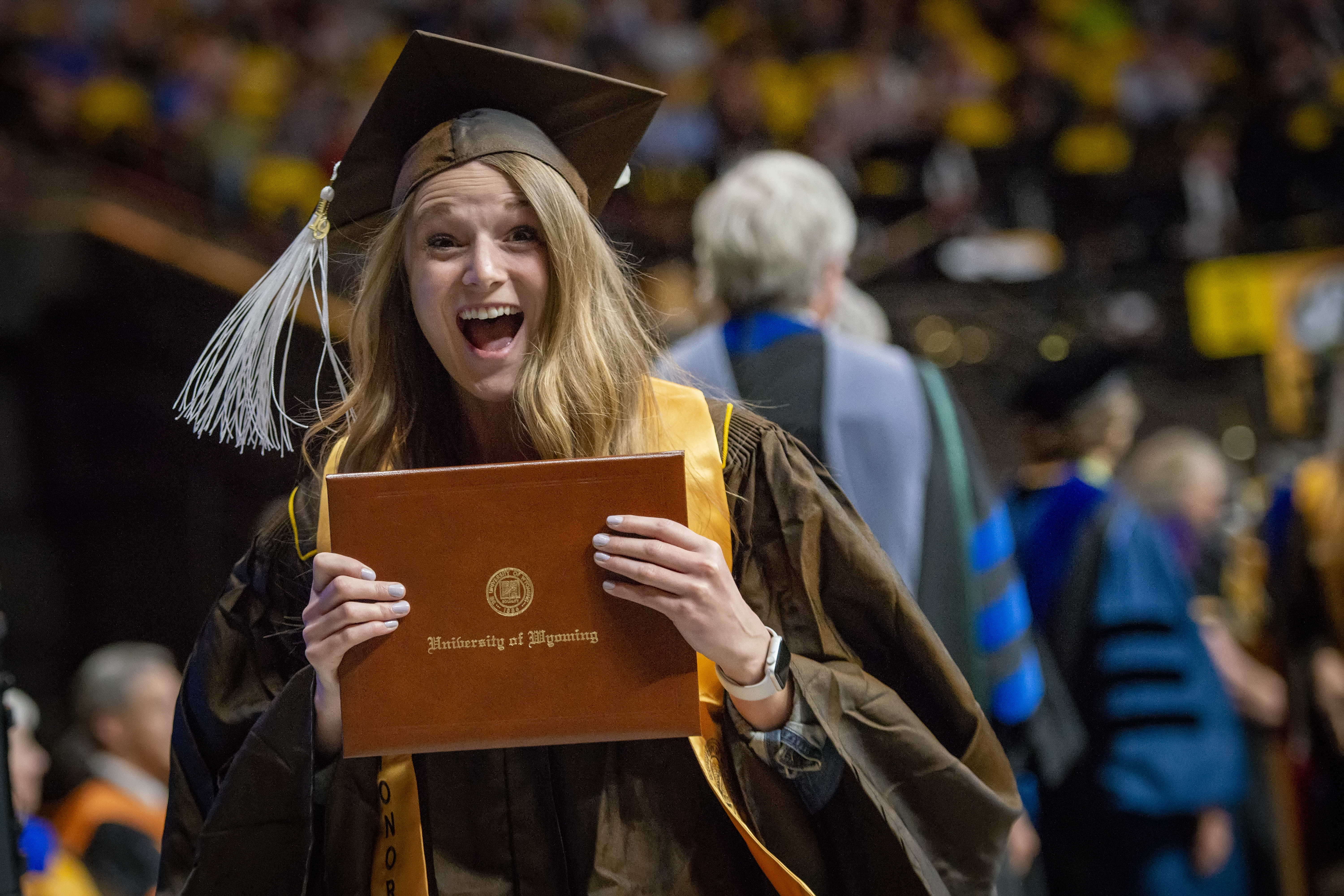 an excited UW student looks very happy while wearing a cap and gown and holding up a diploma.
