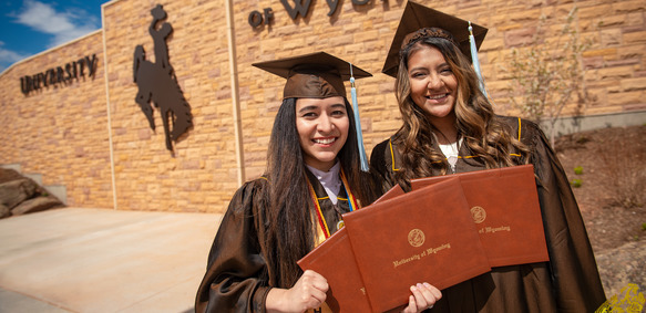 Two UW graduates stand and smile broadly with their degree in front of a University of Wyoming building.