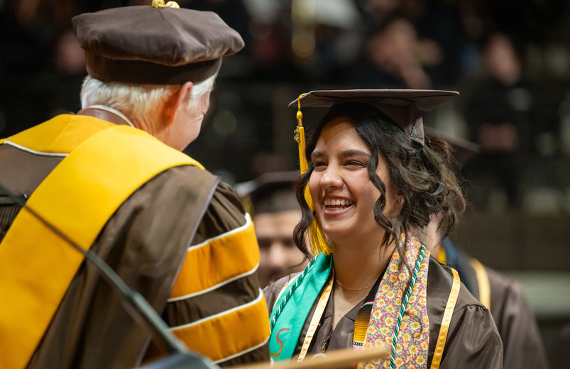A UW transfer student wears a graduation cap and gown and smiles as they shake hands with a UW faculty member during a University of Wyoming graduation ceremony.