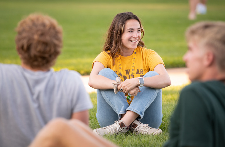 A student sits in the grass wearing a yellow shirt and jeans.