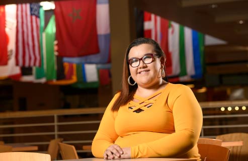 A student sits at a table with international flags in the background.