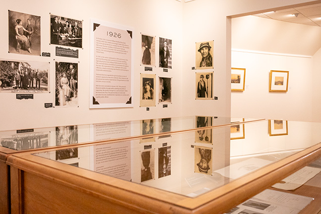 Scene from an exhibit hall. In the foreground is a glass case with documents and other print materials. In the background is a large panel with the headline "1926." Black and white photos of Nellie Tayloe Ross are on display on the wall.