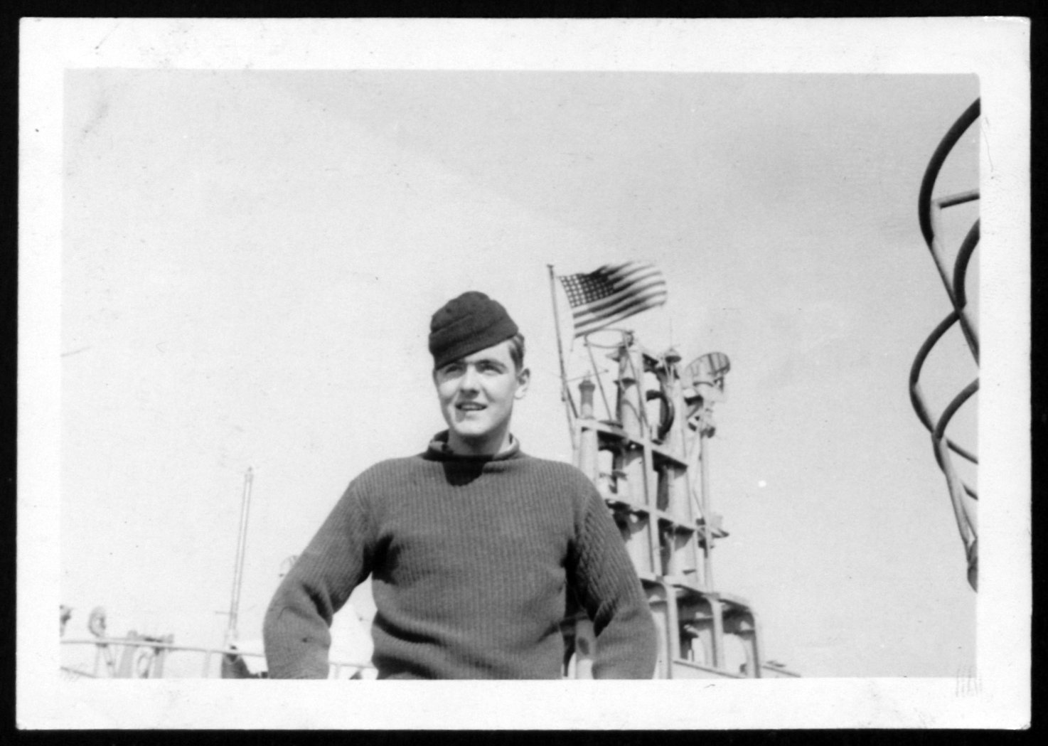 A sailor poses on the deck of a ship. An american flag can be seen in the background