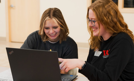 Two students work at a table with a laptop in front of them as they study the screen with smiles on their faces.